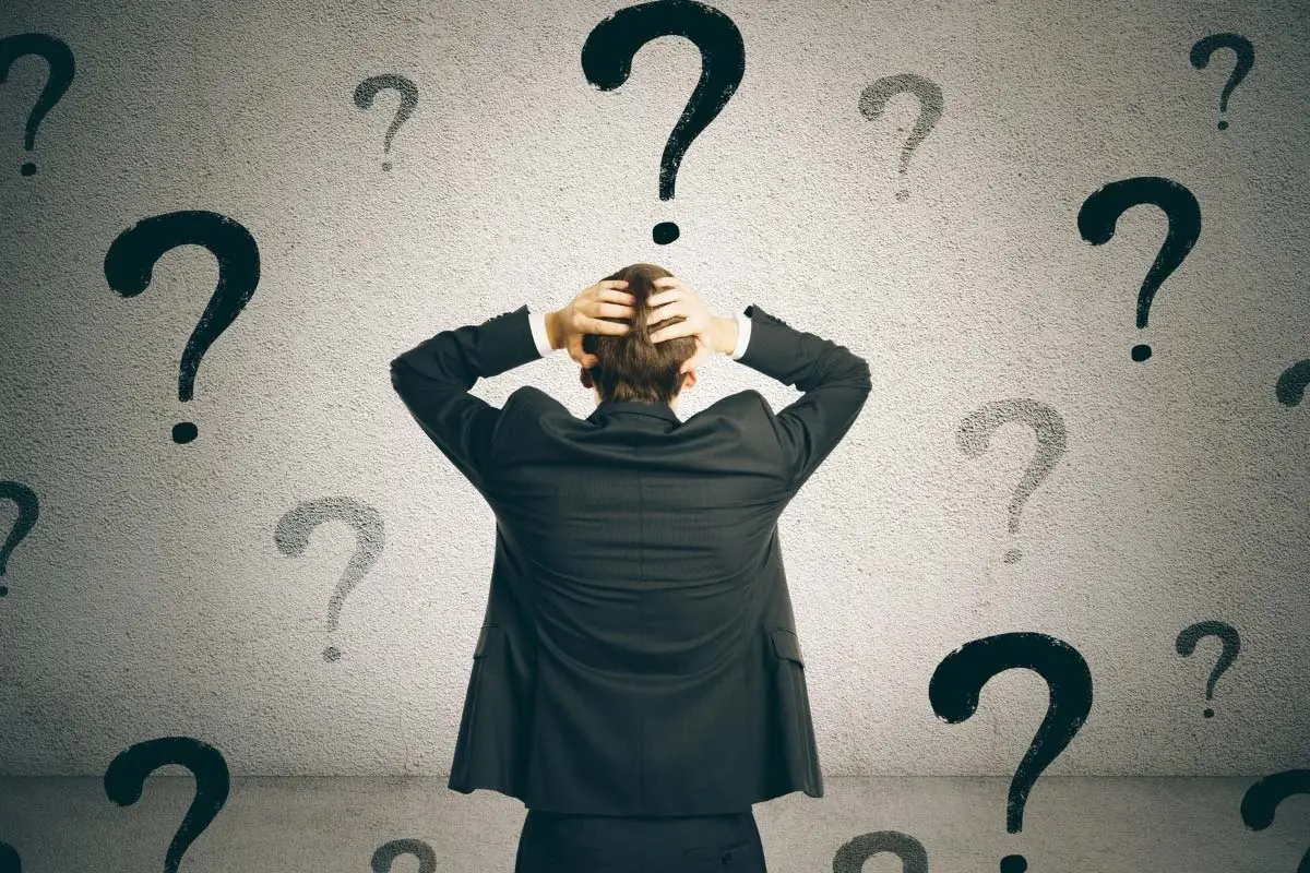 Man in a suit facing a wall covered in large question marks with his hands on his head in frustration.